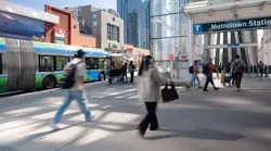 Pedestrians cross a plaza outside Metrotown Station in Burnaby, with a TransLink articulated bus passing behind them and the SkyTrain platform's escalators visible under a glass canopy, surrounded by high-rise buildings.