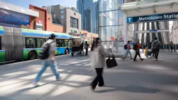 Pedestrians cross a plaza outside Metrotown Station in Burnaby, with a TransLink articulated bus passing behind them and the SkyTrain platform's escalators visible under a glass canopy, surrounded by high-rise buildings.