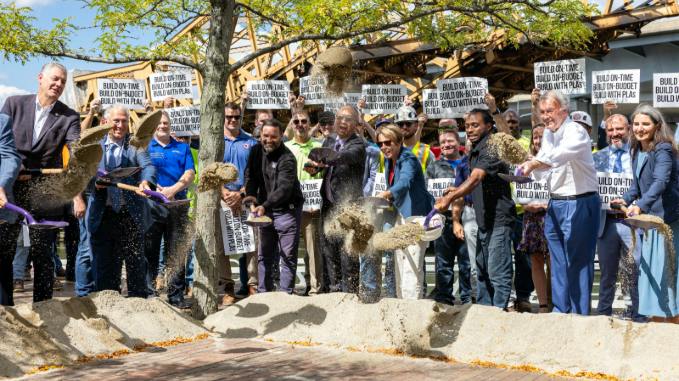 A large group of officials, construction workers and stakeholders simultaneously toss shovels of dirt at a ceremonial groundbreaking event, with crowd members holding 'Build On-Time, Build On-Budget, Build With PLAs' signs in the background.