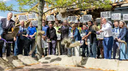 A large group of officials, construction workers and stakeholders simultaneously toss shovels of dirt at a ceremonial groundbreaking event, with crowd members holding 'Build On-Time, Build On-Budget, Build With PLAs' signs in the background.