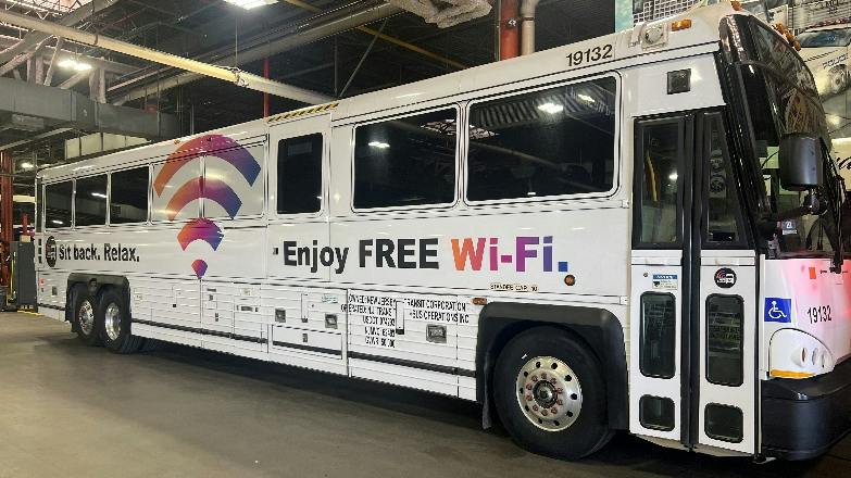 A white NJ Transit motorcoach, numbered 19132, parked inside a maintenance garage, featuring a 'Sit back. Relax. Enjoy FREE Wi-Fi.' ad wrap along its side.