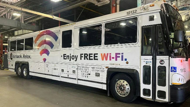 A white NJ Transit motorcoach, numbered 19132, parked inside a maintenance garage, featuring a 'Sit back. Relax. Enjoy FREE Wi-Fi.' ad wrap along its side.