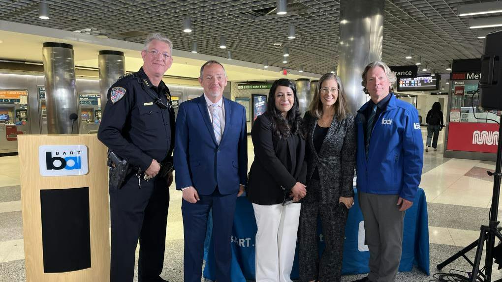 Five BART officials and law enforcement representatives pose for a photo at a press event inside Powell Street Station in San Francisco, standing beside a BART-branded podium and display table, with Muni signage and fare gates visible in the background.