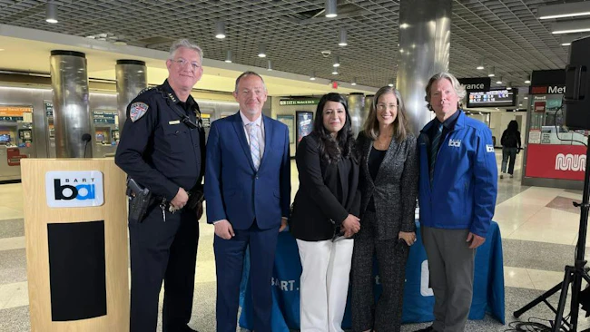 Five BART officials and law enforcement representatives pose for a photo at a press event inside Powell Street Station in San Francisco, standing beside a BART-branded podium and display table, with Muni signage and fare gates visible in the background.