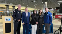 Five BART officials and law enforcement representatives pose for a photo at a press event inside Powell Street Station in San Francisco, standing beside a BART-branded podium and display table, with Muni signage and fare gates visible in the background.