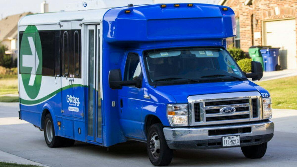 A blue and white GoTaps paratransit shuttle van, numbered 325, drives down a residential street past brick homes and green lawns.