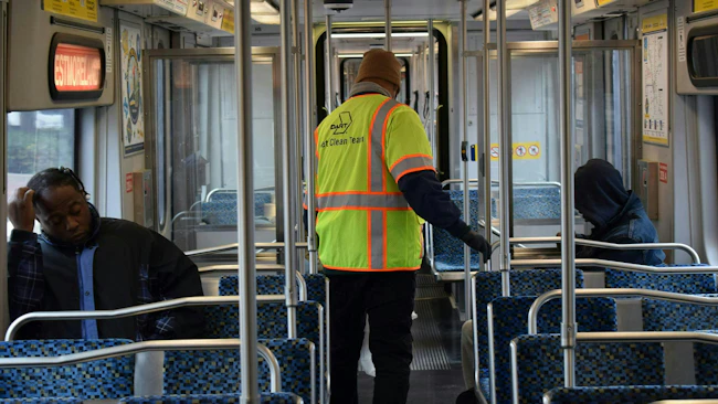 A DART Fleet Clean Team worker in a yellow and orange high-visibility safety vest moves through the interior of a DART light-rail car, with blue patterned seating and two riders visible on either side of the aisle.