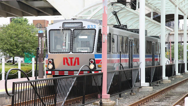 A Greater Cleveland RTA light rail vehicle, numbered 833 and signed 'Waterfront,' stops at a covered platform station, with a tree-lined area and parked cars visible in the background.