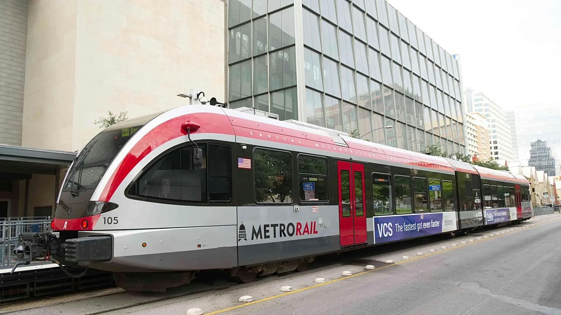 Capital Metro MetroRail diesel multiple unit car number 105 in a white and red livery sits at a downtown Austin station platform, with the Austin skyline and modern office buildings visible in the background under an overcast sky.