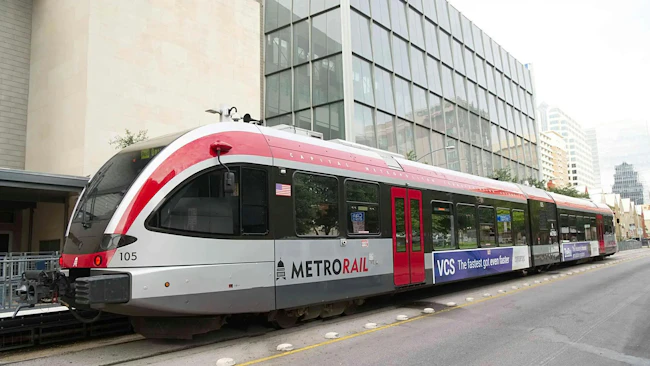 Capital Metro MetroRail diesel multiple unit car number 105 in a white and red livery sits at a downtown Austin station platform, with the Austin skyline and modern office buildings visible in the background under an overcast sky.
