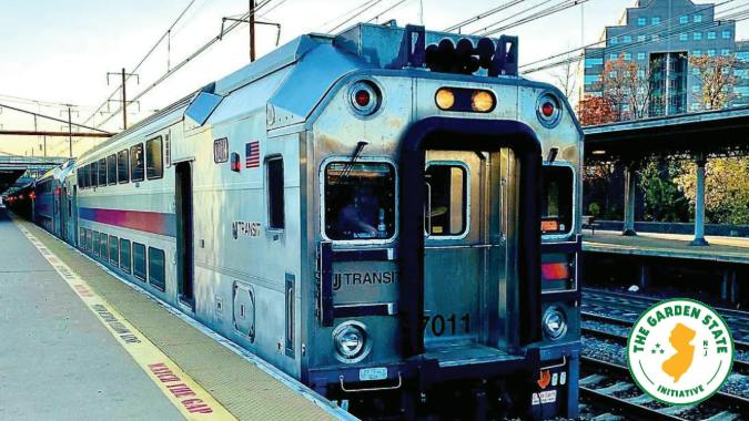 An NJ Transit bi-level rail car, numbered 7011, sits at a platform beside a 'Mind the Gap' warning strip, with the Garden State Initiative logo overlaid in the corner.