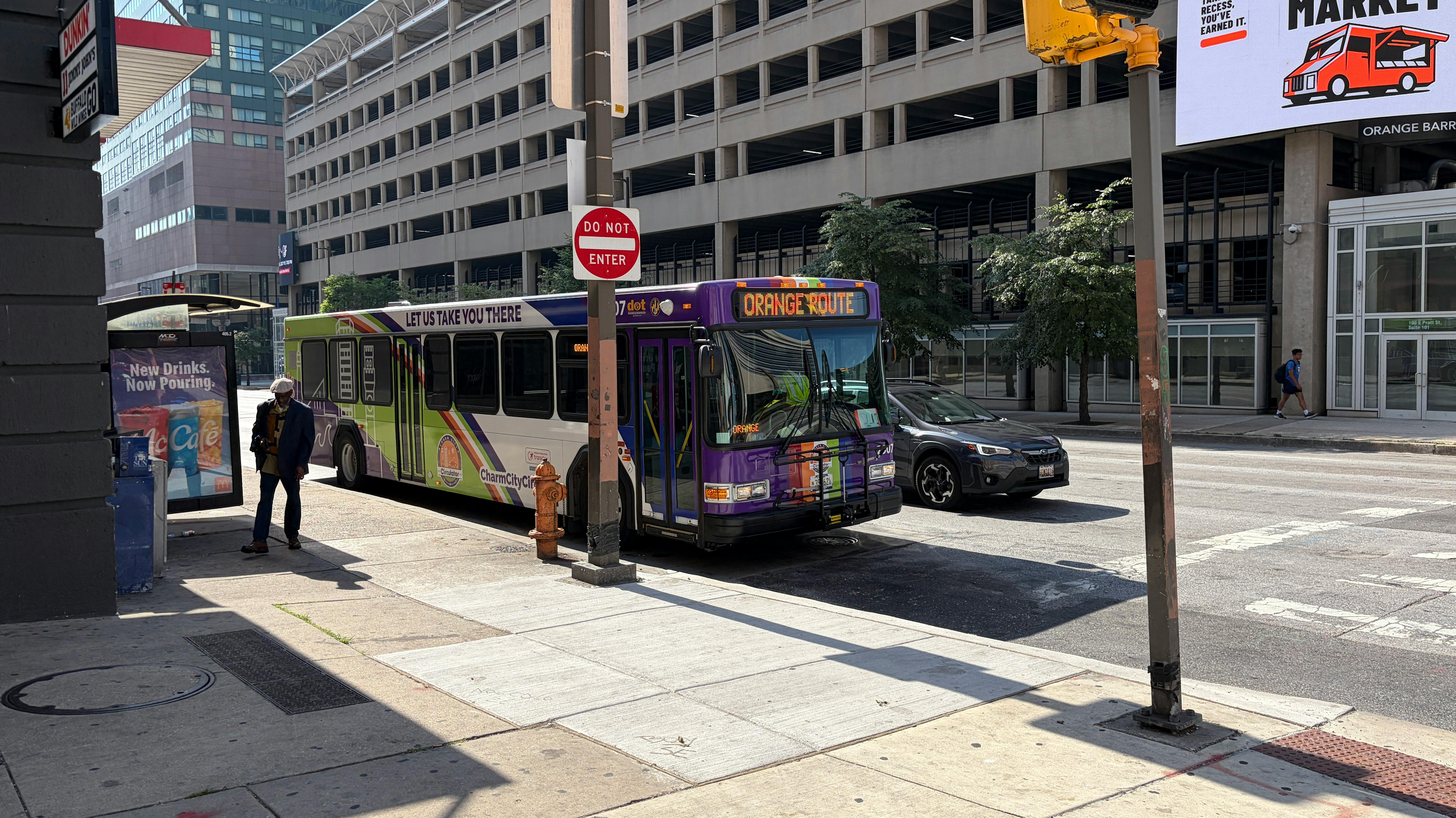 A Charm City Circulator bus signed 'Orange Route' turns onto a downtown Baltimore street, passing a bus shelter and pedestrian as a parking garage and storefronts line the block.