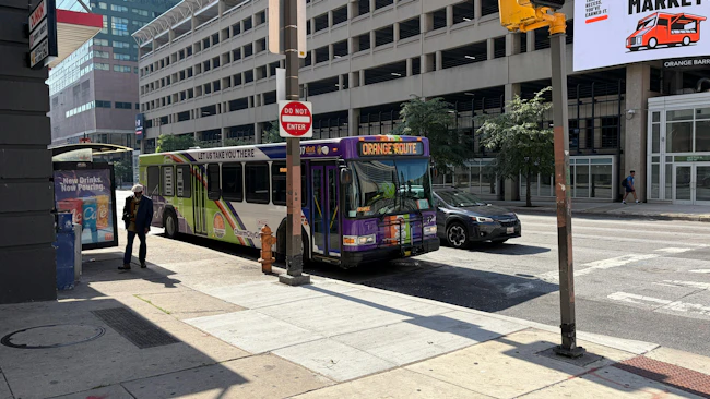 A Charm City Circulator bus signed 'Orange Route' turns onto a downtown Baltimore street, passing a bus shelter and pedestrian as a parking garage and storefronts line the block.