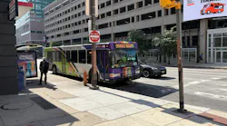 A Charm City Circulator bus signed 'Orange Route' turns onto a downtown Baltimore street, passing a bus shelter and pedestrian as a parking garage and storefronts line the block.