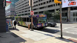 A Charm City Circulator bus signed 'Orange Route' turns onto a downtown Baltimore street, passing a bus shelter and pedestrian as a parking garage and storefronts line the block.