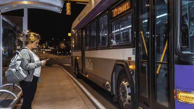 A passenger checks her phone while waiting under a covered shelter as a WeGo Public Transit bus, signed for Route 17, arrives at night.