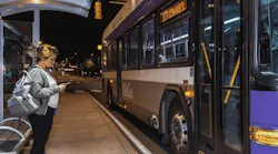 A passenger checks her phone while waiting under a covered shelter as a WeGo Public Transit bus, signed for Route 17, arrives at night.