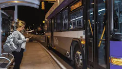 A passenger checks her phone while waiting under a covered shelter as a WeGo Public Transit bus, signed for Route 17, arrives at night.