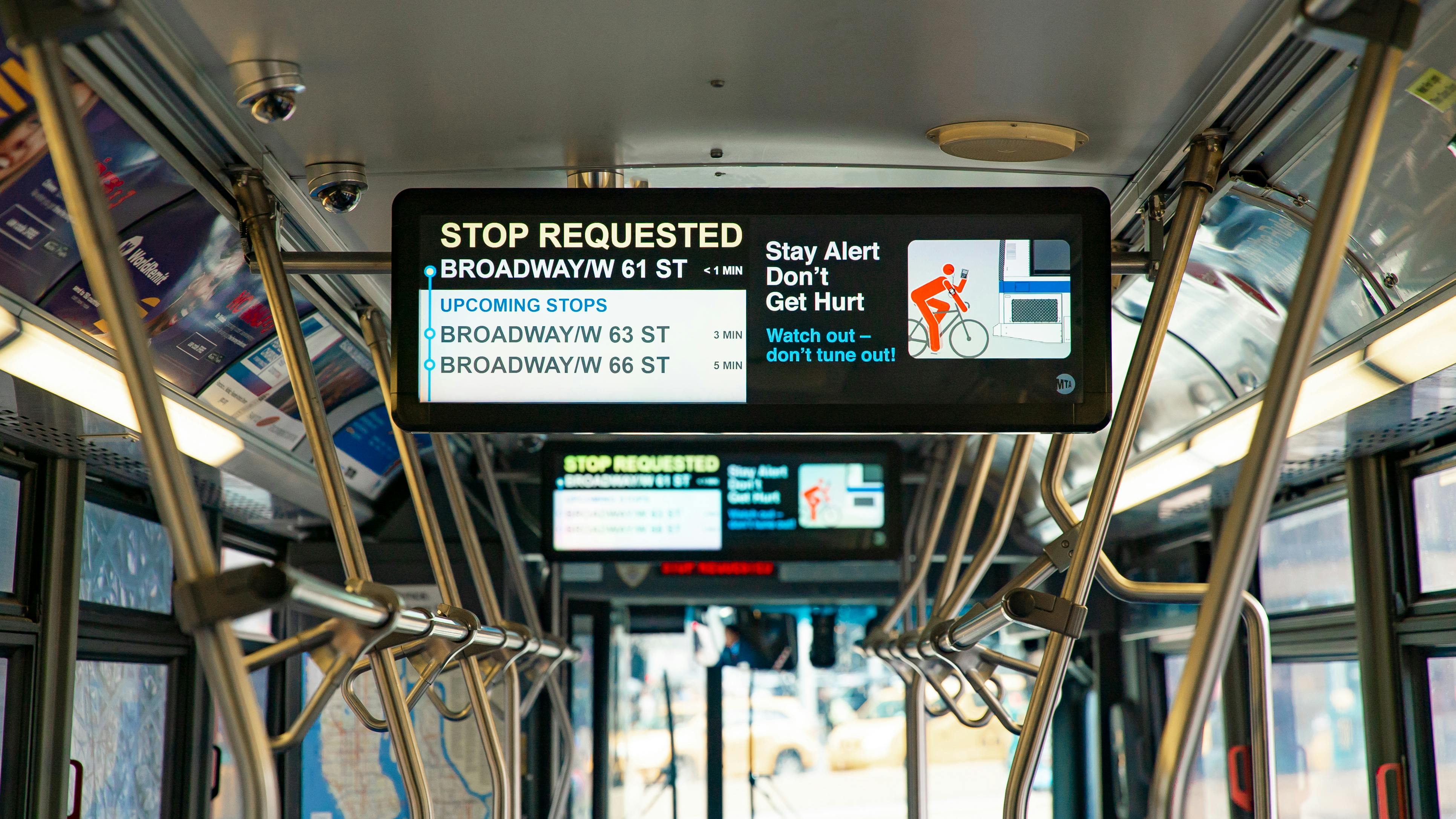 Interior view of an MTA New York City Bus showing a Hitachi Mobility CleverVision passenger information display, indicating 'Stop Requested' for Broadway/W 61 St along with upcoming stops and a bicyclist safety message.