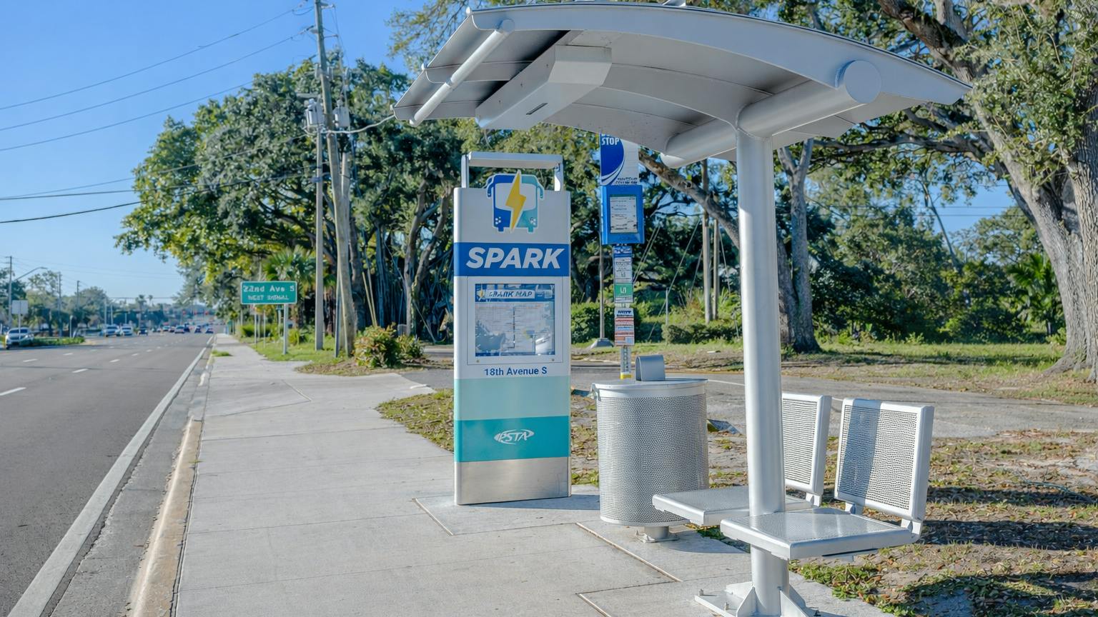A PSTA SPARK bus rapid transit stop on 18th Avenue South, featuring Tolar's branded totem sign with route map and a real-time e-paper passenger information display, alongside a shaded shelter with metal benches along the roadside.