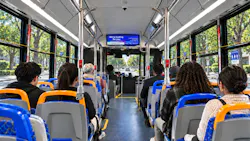 Interior view of a bus showing the ETA Onboard Infotainment Display mounted above the aisle, showing a 'We're looking for you' hiring message alongside the date and time, with seated passengers filling the rows below.