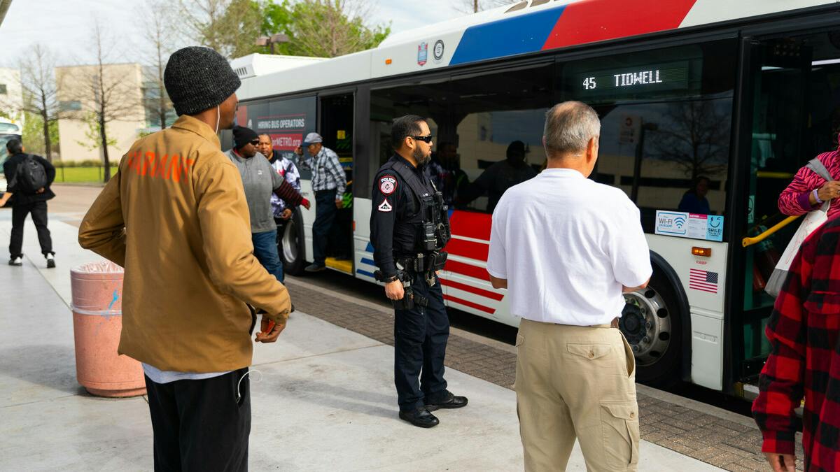A Houston Metropolitan Transit Authority police officer stands watch as riders board a METRO bus displaying Route 45 Tidwell on its destination board at a transit center, with the agency's red, white and blue livery and a 'Hiring Bus Operators' advertisement visible on the bus exterior.