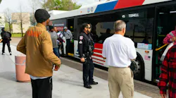 A Houston Metropolitan Transit Authority police officer stands watch as riders board a METRO bus displaying Route 45 Tidwell on its destination board at a transit center, with the agency's red, white and blue livery and a 'Hiring Bus Operators' advertisement visible on the bus exterior.