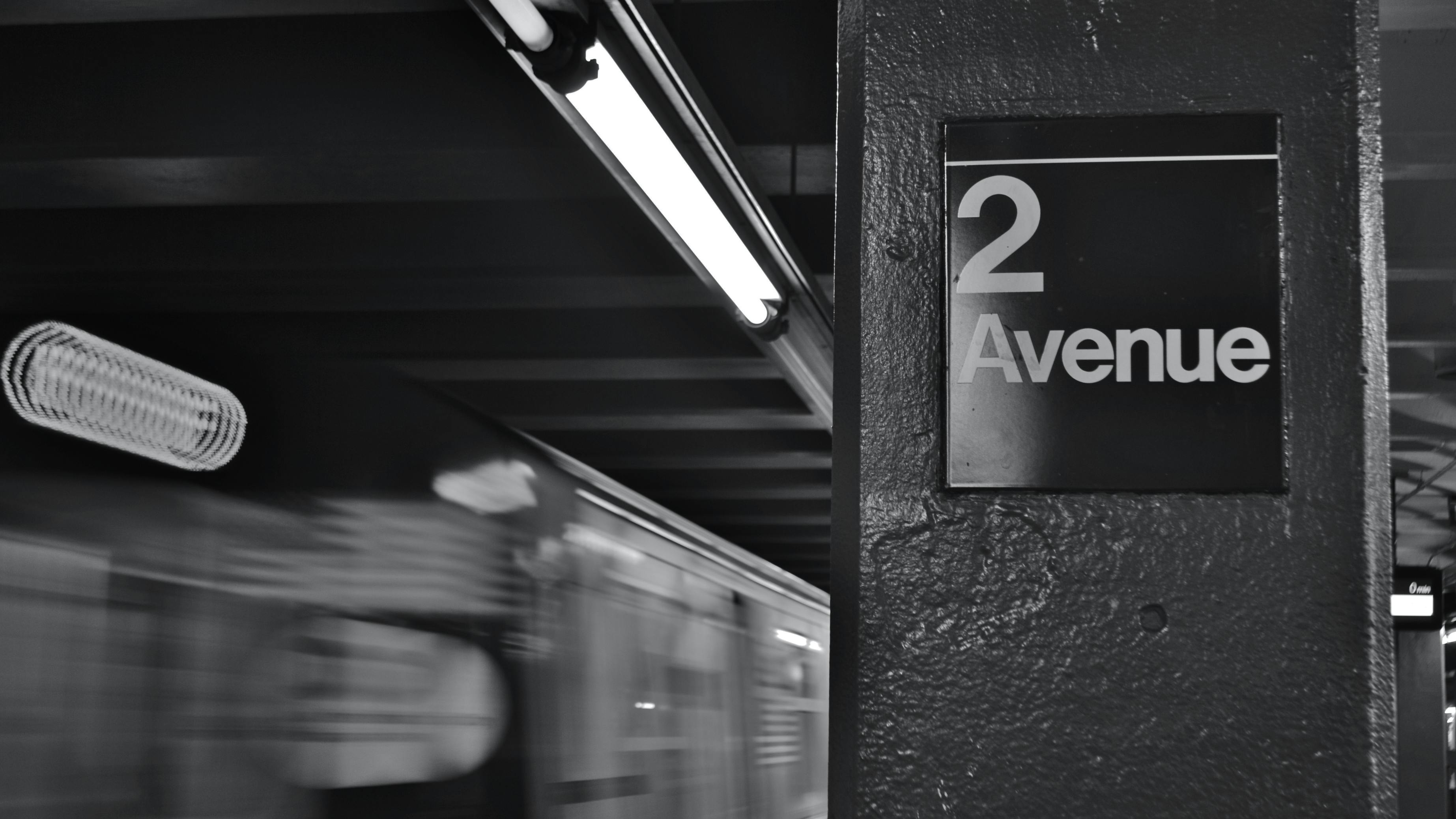 Black-and-white long-exposure photo of a subway train passing the '2 Avenue' station sign, with motion blur from the train and overhead fluorescent lighting.