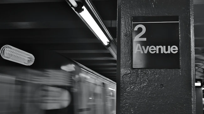 Black-and-white long-exposure photo of a subway train passing the '2 Avenue' station sign, with motion blur from the train and overhead fluorescent lighting.