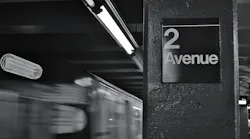 Black-and-white long-exposure photo of a subway train passing the '2 Avenue' station sign, with motion blur from the train and overhead fluorescent lighting.