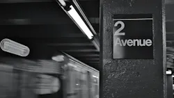 Black-and-white long-exposure photo of a subway train passing the '2 Avenue' station sign, with motion blur from the train and overhead fluorescent lighting.