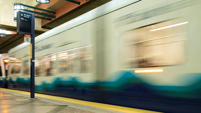 Long-exposure photo of a Sound Transit Link light rail train, in blue and white livery with a wave graphic, pulling out of a station past a 'Bus Bay D' wayfinding sign for Route 550 to Bellevue.