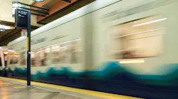 Long-exposure photo of a Sound Transit Link light rail train, in blue and white livery with a wave graphic, pulling out of a station past a 'Bus Bay D' wayfinding sign for Route 550 to Bellevue.