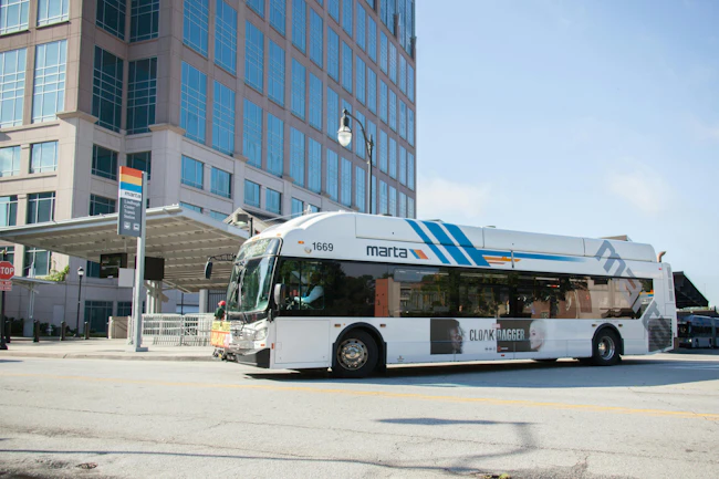 MARTA bus number 1669 in the agency's white livery with blue and orange diagonal stripes departs the Lindbergh Center Transit Station in Atlanta, carrying a Marvel's Cloak & Dagger advertisement wrap on its lower body, with a MARTA bus stop sign and modern office tower visible in the background.