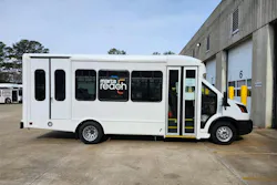 A white Ford Transit-based MARTA Reach on-demand microtransit van with the 'marta reach' logo displayed on its passenger windows, parked outside a transit maintenance facility in Atlanta, with additional MARTA buses visible in the background