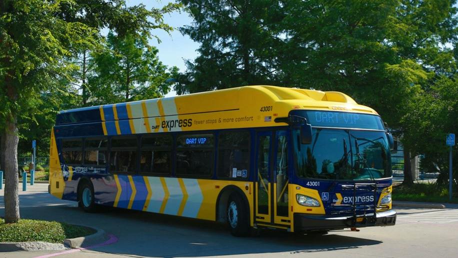 A yellow and blue DART Express bus, unit 43001, displays a 'DART VIP' destination sign while parked near trees on a paved lot.