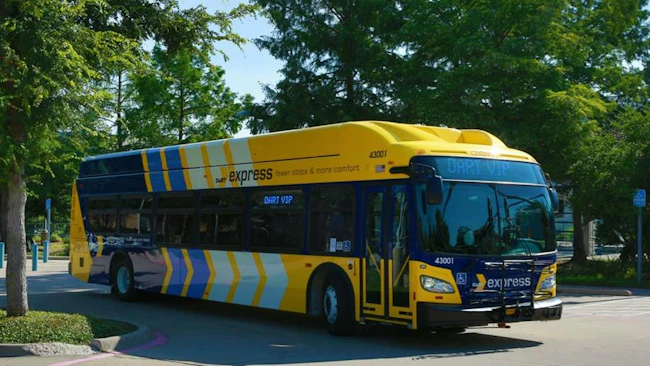 A yellow and blue DART Express bus, unit 43001, displays a 'DART VIP' destination sign while parked near trees on a paved lot.