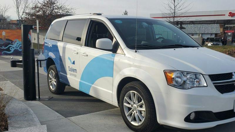 A white Dodge Grand Caravan branded as 'Salem Skipper' with blue wave graphics is parked at a curb near a charging station and gas station in the background.