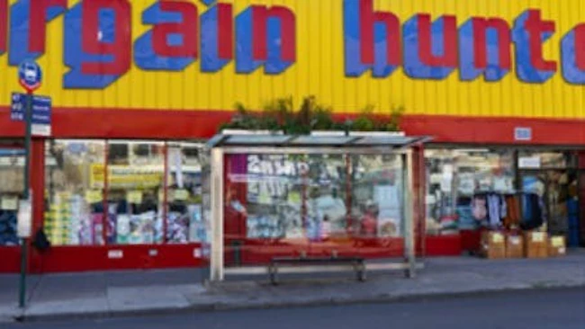 A glass-enclosed bus shelter with a bench and a green roof planted with vegetation sits on a New York City sidewalk in Brownsville, in front of a storefront reading 'Bargain Hunt.'