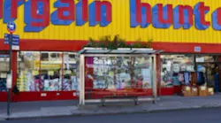 A glass-enclosed bus shelter with a bench and a green roof planted with vegetation sits on a New York City sidewalk in Brownsville, in front of a storefront reading 'Bargain Hunt.'