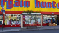 A glass-enclosed bus shelter with a bench and a green roof planted with vegetation sits on a New York City sidewalk in Brownsville, in front of a storefront reading 'Bargain Hunt.'