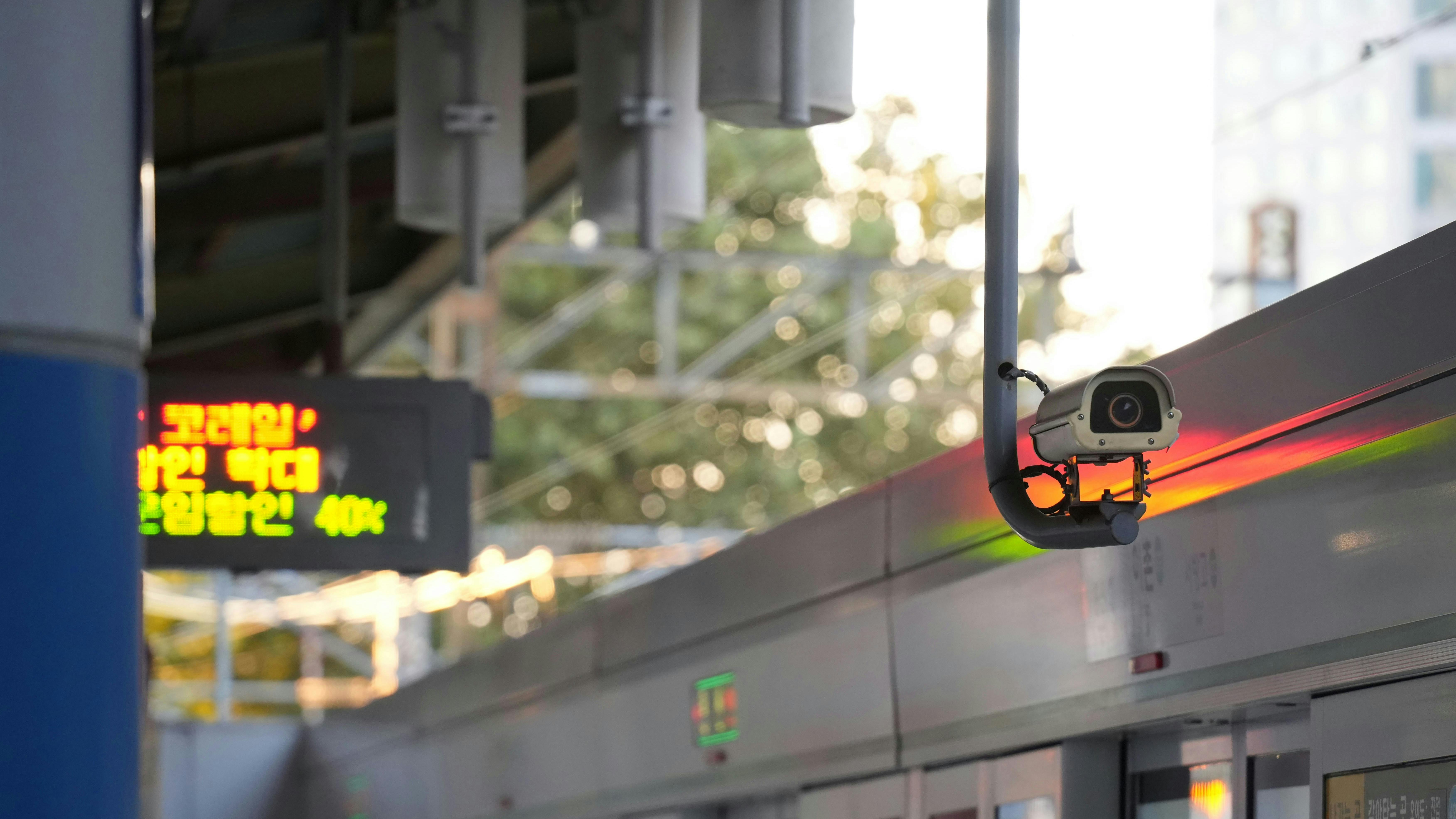 A security camera mounted above a train door at a South Korean transit platform, with a Korean-language LED display and sunlit trees visible in the background.