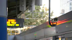 A security camera mounted above a train door at a South Korean transit platform, with a Korean-language LED display and sunlit trees visible in the background.