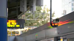 A security camera mounted above a train door at a South Korean transit platform, with a Korean-language LED display and sunlit trees visible in the background.