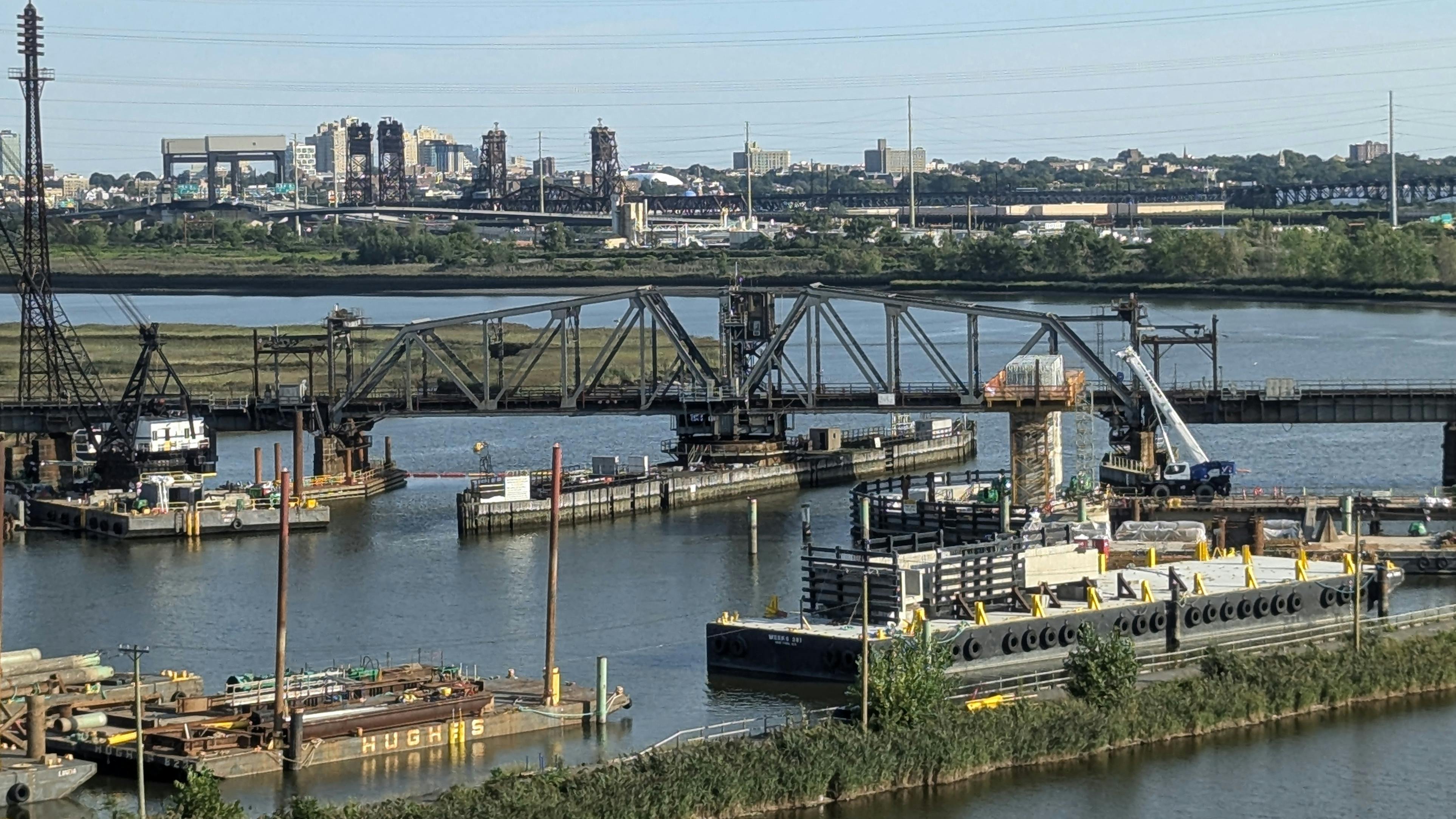 A representative photo of a rail bridge and surrounding waterway construction site, used to illustrate the Portal North Bridge project's second cutover for its final track, with barges, cranes, and the Newark, New Jersey skyline visible in the background.