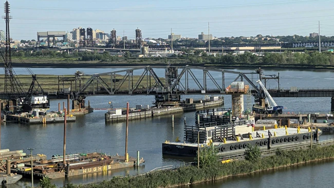 A representative photo of a rail bridge and surrounding waterway construction site, used to illustrate the Portal North Bridge project's second cutover for its final track, with barges, cranes, and the Newark, New Jersey skyline visible in the background.