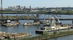 A representative photo of a rail bridge and surrounding waterway construction site, used to illustrate the Portal North Bridge project's second cutover for its final track, with barges, cranes, and the Newark, New Jersey skyline visible in the background.