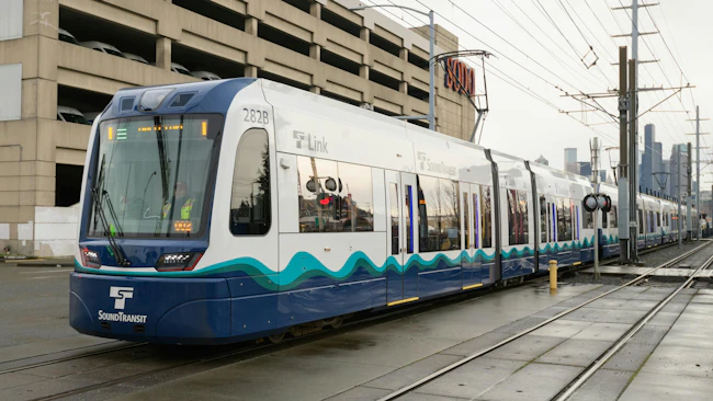 A Sound Transit Link light-rail vehicle, car 282B, travels on tracks past the SODO neighborhood in Seattle, with the city skyline visible in the background.