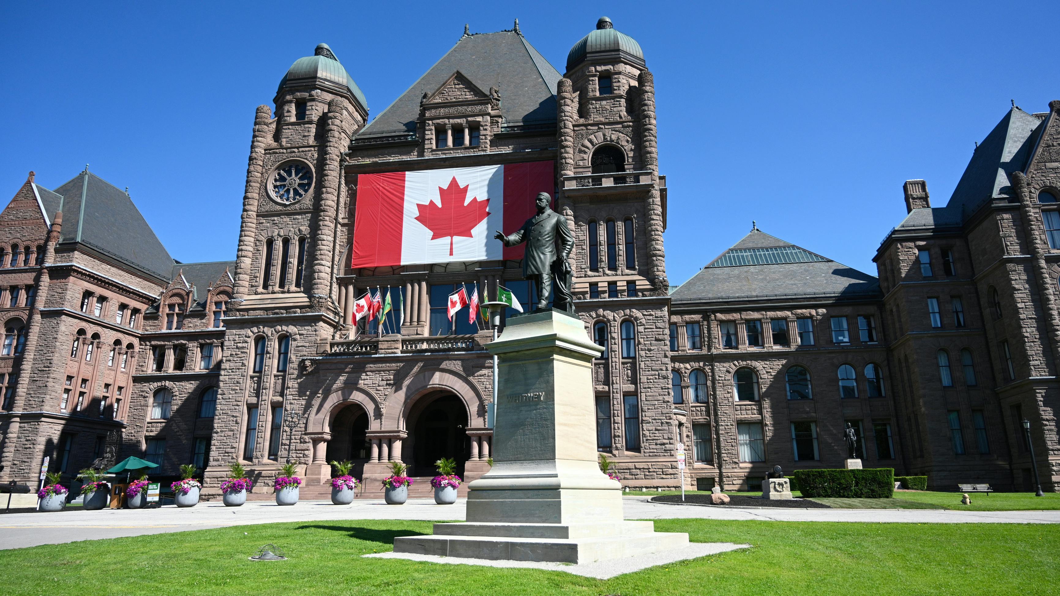The Ontario Legislative Building at Queen's Park in Toronto, its sandstone facade draped with a large Canadian flag, with a statue of former premier Whitney in the foreground and flowering planters lining the entrance.