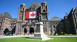 The Ontario Legislative Building at Queen's Park in Toronto, its sandstone facade draped with a large Canadian flag, with a statue of former premier Whitney in the foreground and flowering planters lining the entrance.